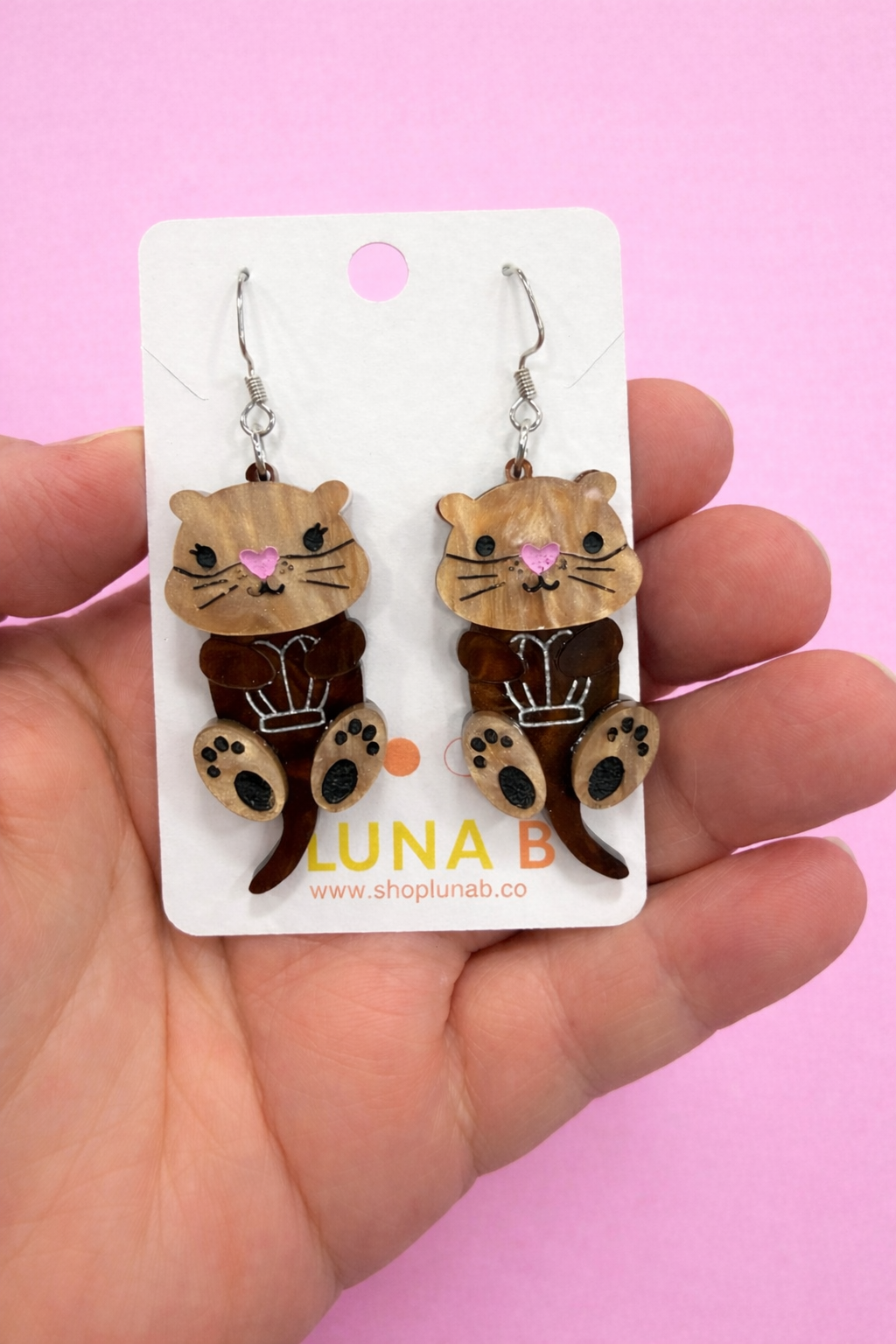 Otter shaped earrings on a Luna B branded card held by a hand against a pink background