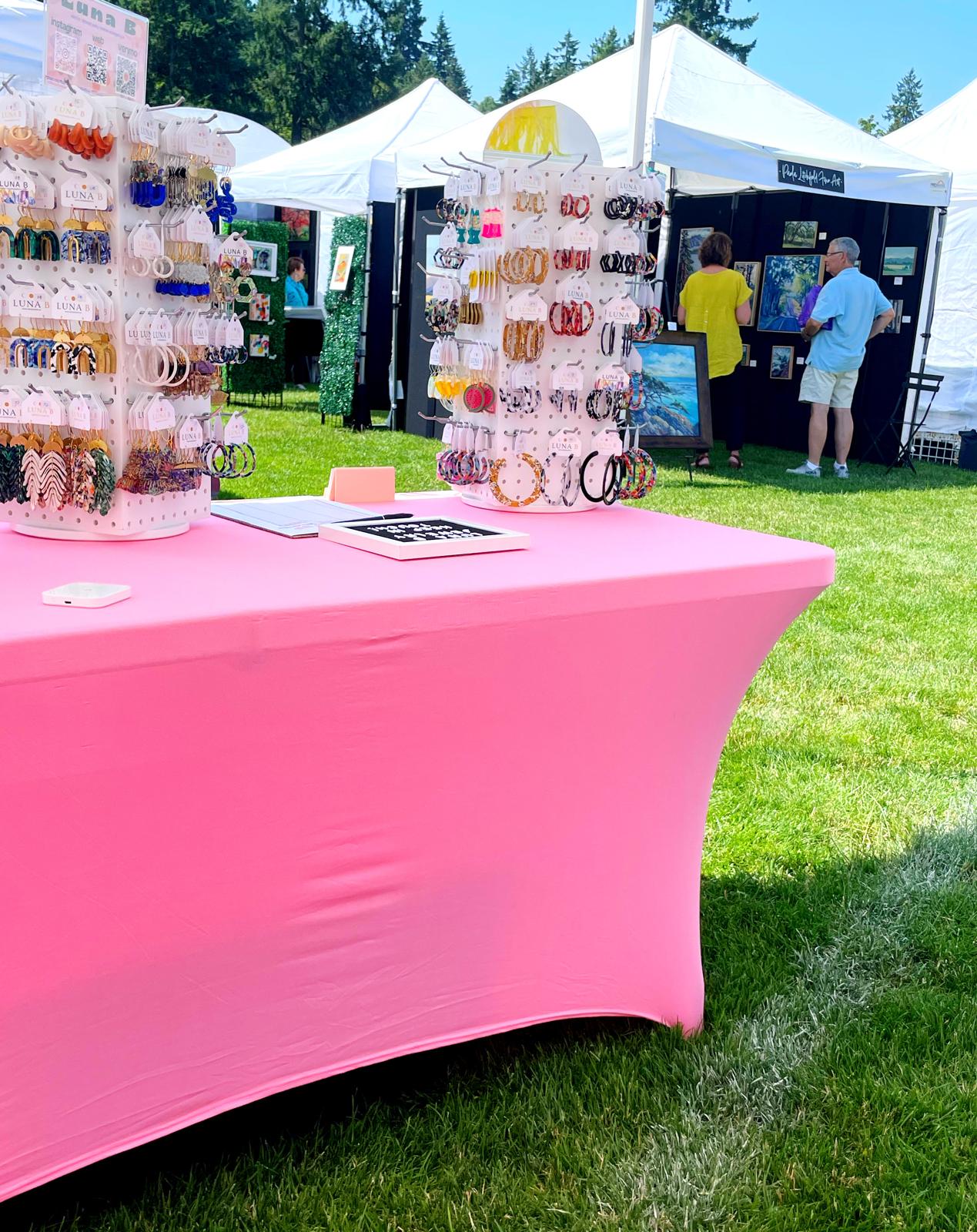 Table on grass with a pink table cloth, with white jewelry spinner displays on top. In the background there are art tents at an art festival.