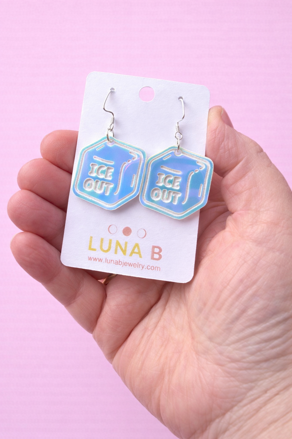 Blue 'ICE OUT' earrings on a white card held by a hand against a pink background
