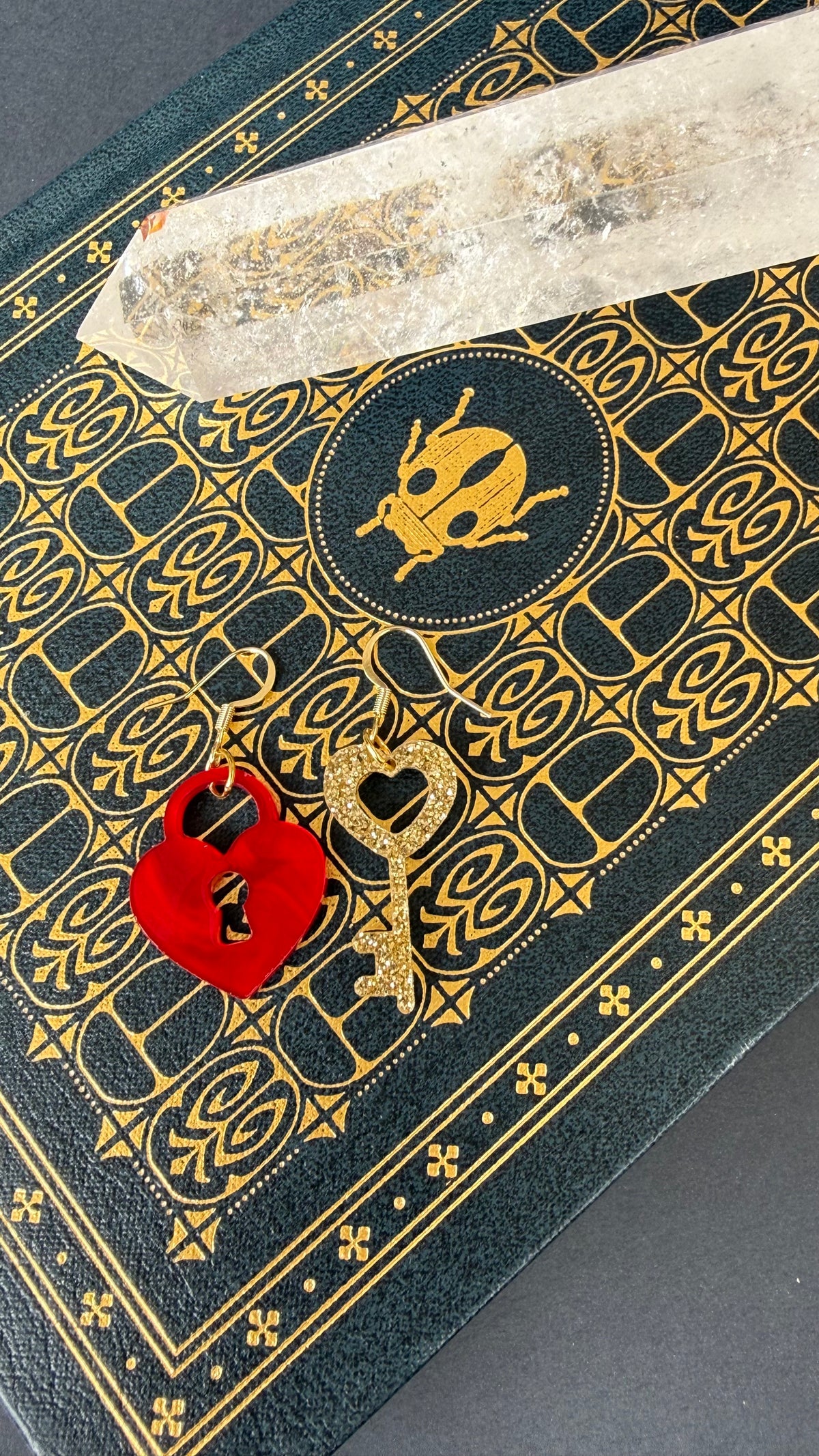 A close-up of a decorative black journal with gold embossed patterns and a skull-and-crossbones emblem, featuring a red wax seal with a keyhole and a gold heart-shaped key resting on top, alongside a clear quartz crystal.