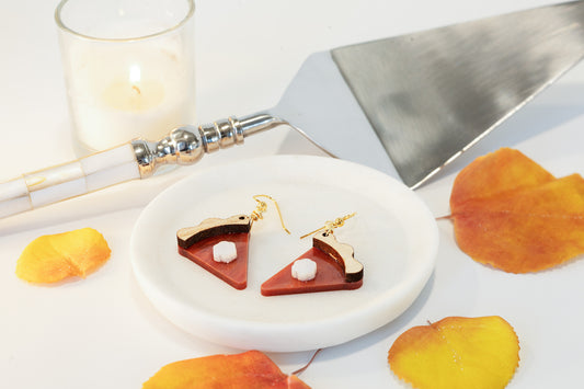 A pair of pumpkin pie-shaped earrings made from copper acrylic, displayed on a white plate with a candle and leaf accents.
