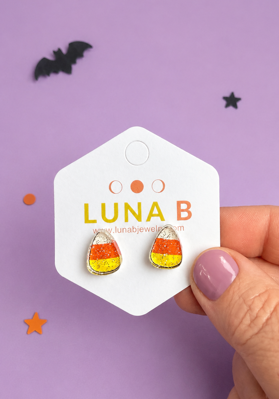 A hand with lavender nail polish holds a white hexagonal card displaying candy corn stud earrings. The card is branded 'LUNA B' with a website, set against a purple background with small black and orange decorative stars and bats.