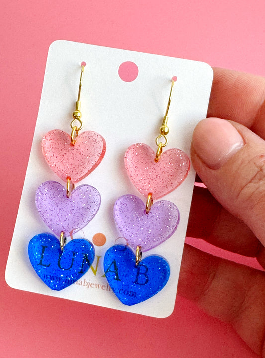 Glittery handmade earrings with stacked pink, purple, and blue acrylic hearts representing the bisexual flag, with gold-plated ear wires.