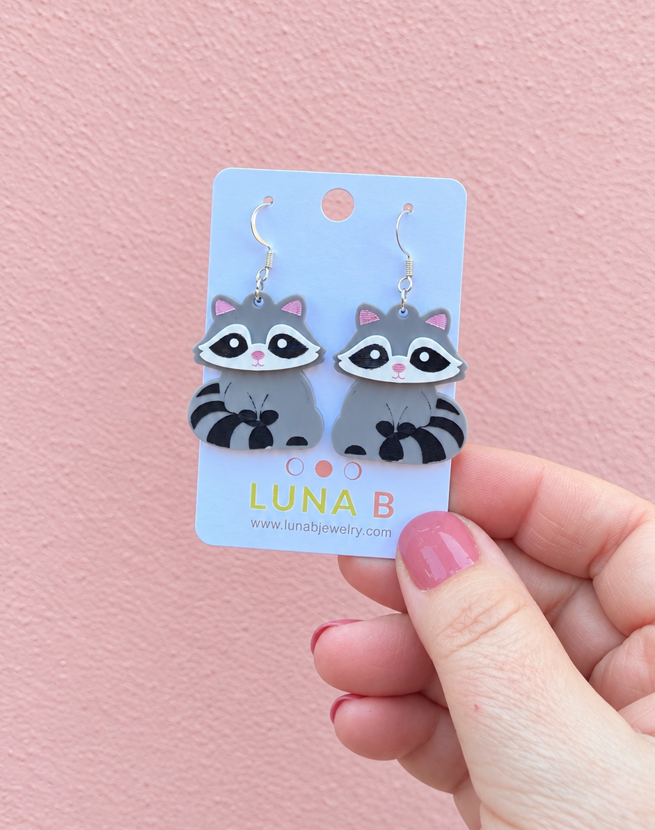 A hand with pink nail polish holds a light blue card displaying a pair of raccoon-shaped dangle earrings against a textured pink wall. The card is branded 'LUNA B'.