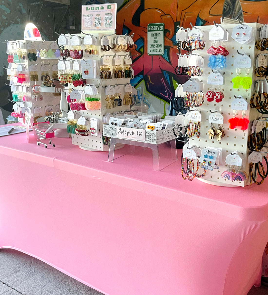a table covered in a pink tablecloth. Three white display spinners sit on top displaying colorful acrylic jewelry for sale.