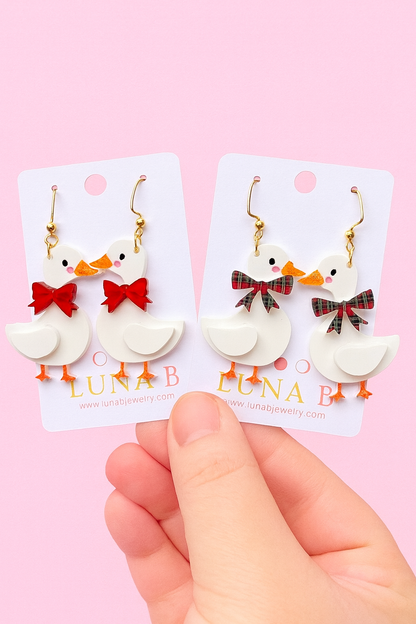 White duck-shaped earrings with red and plaid bows on a Luna B branded card, held against a pink background.