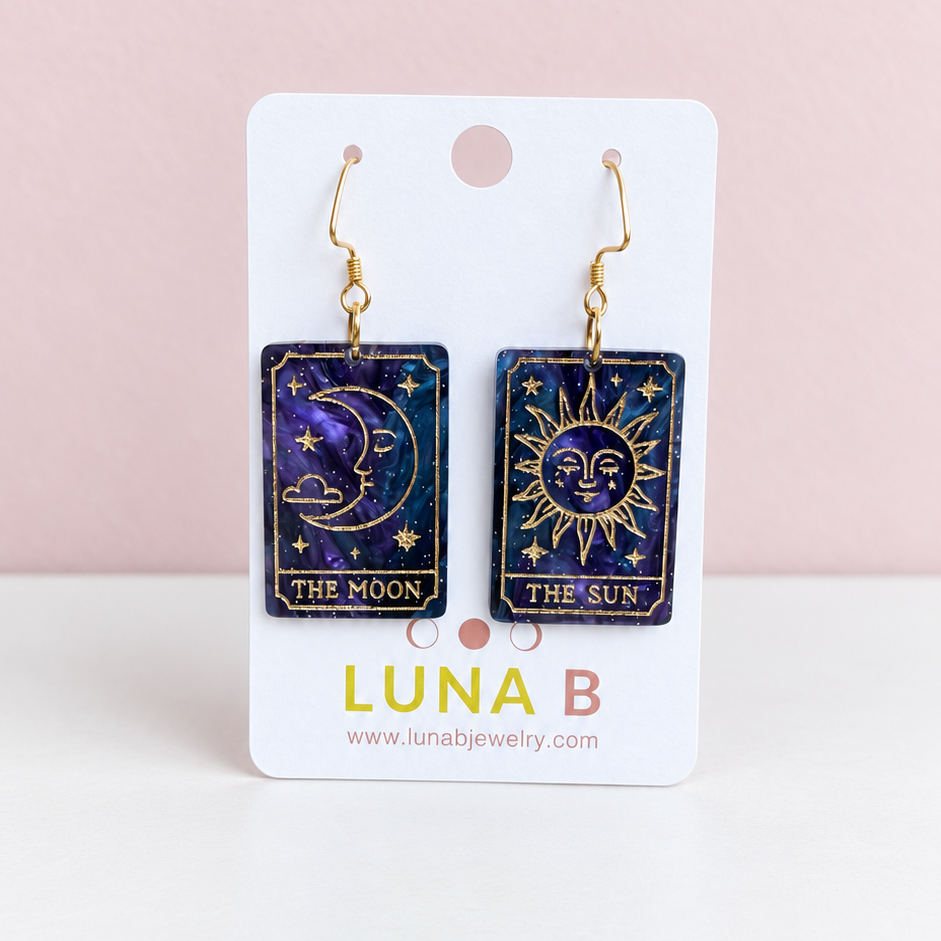 Mismatched Sun and Moon tarot card earrings made from swirling purple and midnight blue acrylic, featuring gold engraved celestial illustrations and gold-tone hooks, displayed on a Luna B Jewelry card against a soft pink background.