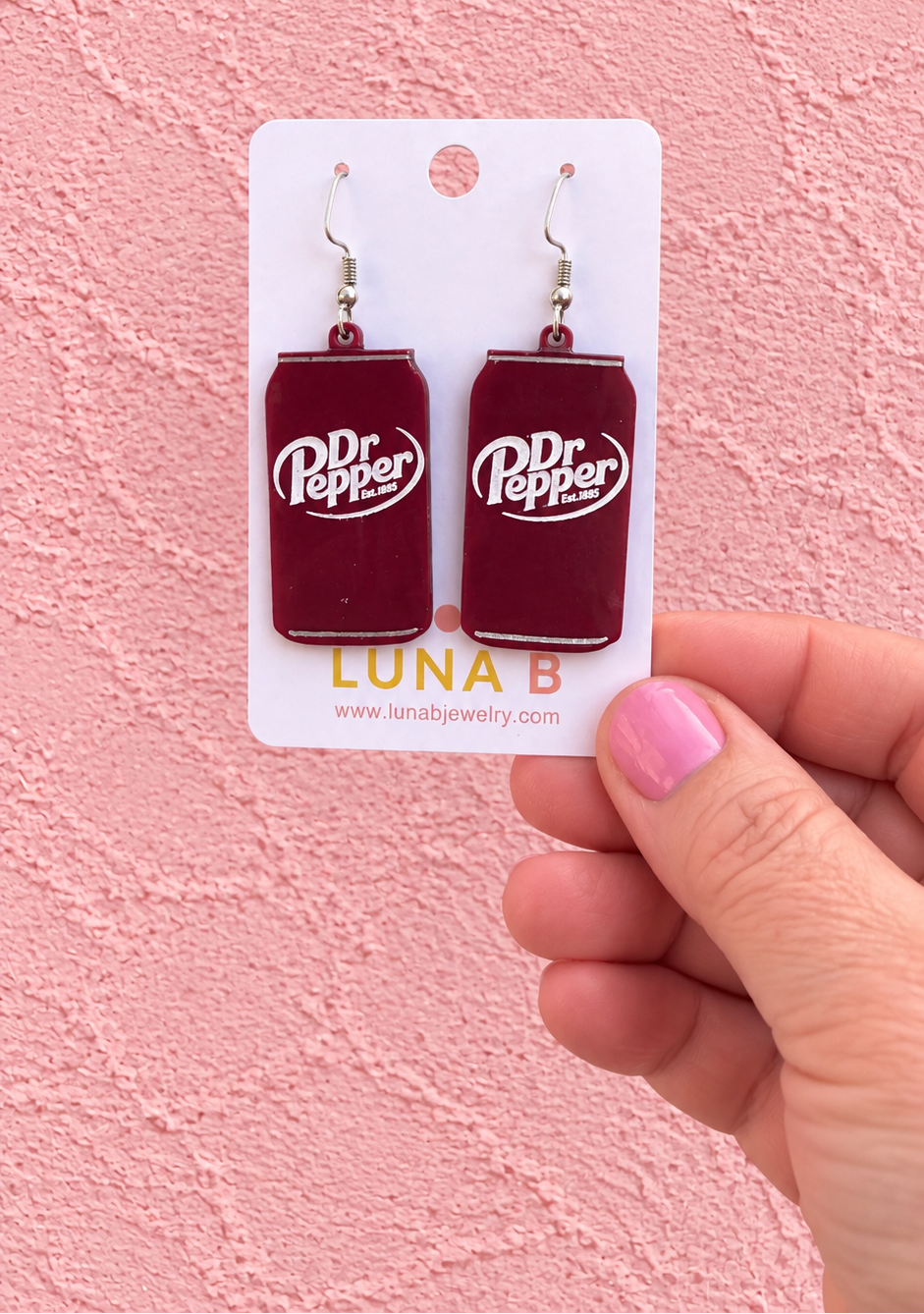 A hand with pink nail polish holds a white card displaying a pair of Dr Pepper can-shaped dangle earrings against a pink textured wall.