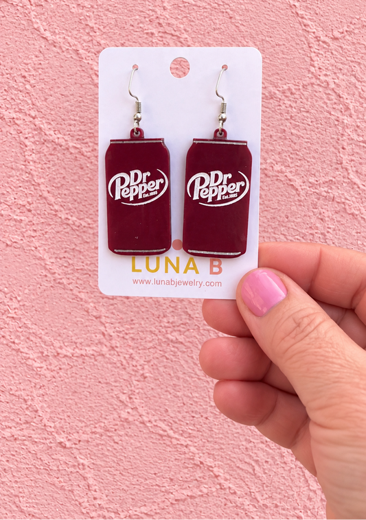 A hand with pink nail polish holds a white card displaying a pair of Dr Pepper can-shaped dangle earrings against a pink textured wall.