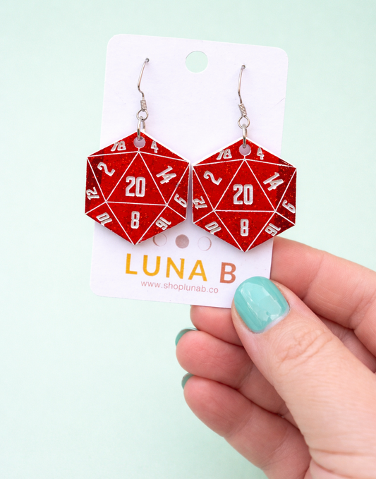 Red geometric 20 sided dice earrings on a white card with 'LUNA B' branding, held by a hand against a light green background.