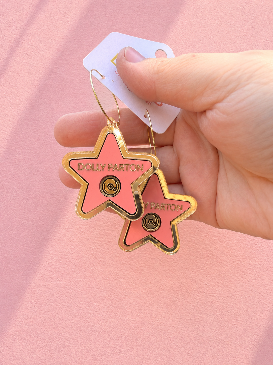 A hand holding a pair of star-shaped gold-tone earrings with pink enamel inlays and the text 'DOLLY PARTON' on a pink background.