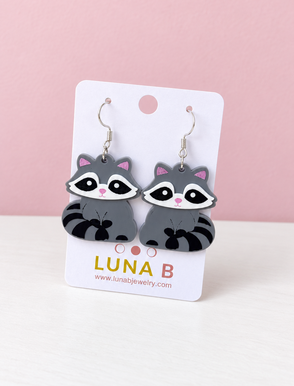 A pair of raccoon-shaped dangle earrings displayed on a white backing card. The earrings feature gray bodies, black and white facial markings, pink inner ears, and silver-tone hooks.