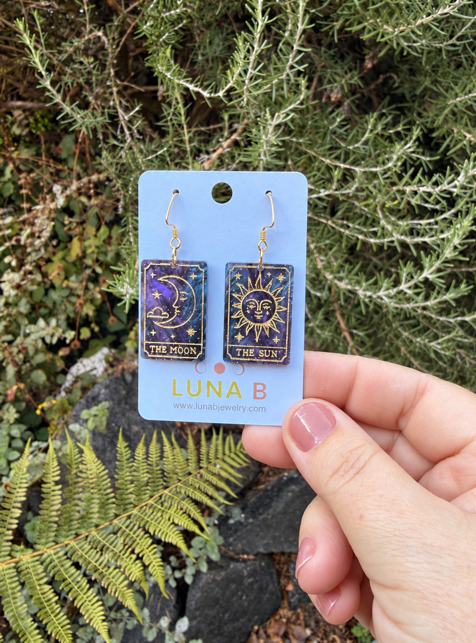 A hand holds a light blue card displaying a pair of tarot card-inspired earrings labeled 'LUNA B'. The earrings feature gold hooks and rectangular panels with celestial designs, including a crescent moon and stars on one side and a sun face with rays on the other.