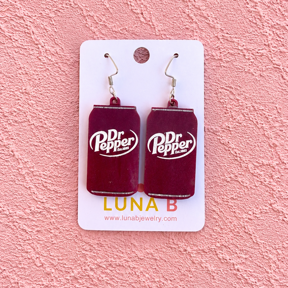 A pair of rectangular dark red earrings with white Dr Pepper logos, displayed on a white backing card against a textured pink background.