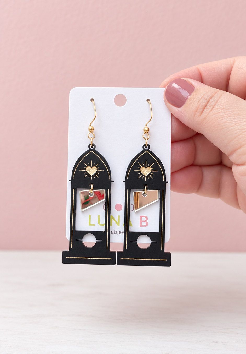 Black acrylic guillotine earrings with pointed Gothic cathedral arches, gold sacred heart accents, and dangling silver mirror blades, displayed on a Luna B Jewelry card against a soft pink background.