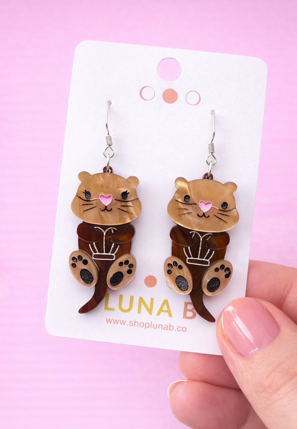 Otter-shaped earrings on a Luna B card against a pink background