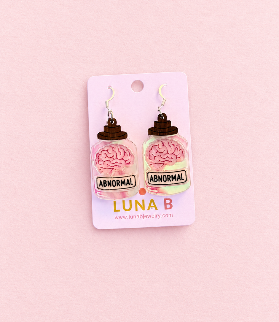 A pair of novelty earrings shaped like jars containing pink brains, mounted on a pink backing card labeled 'LUNA B' against a solid pink background.