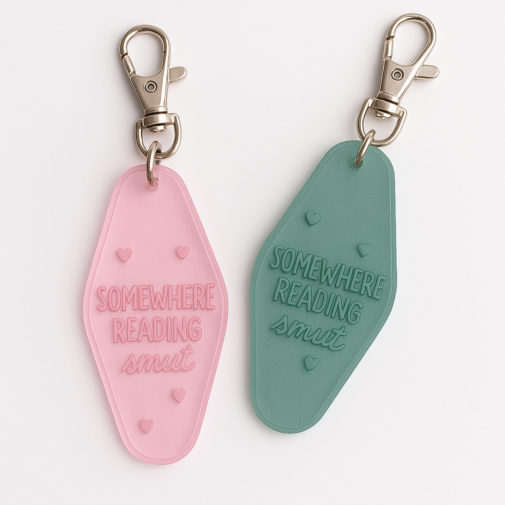 Retro motel-style acrylic keychains in pink and green with engraved text reading ‘Somewhere Reading Smut,’ featuring small heart details and metal clasps.