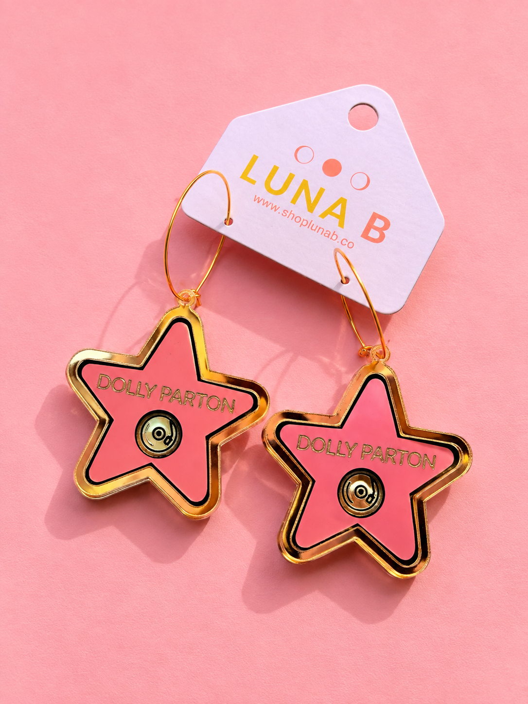 A pair of star-shaped enamel pins attached to a white branded card, photographed on a solid pink background. The pins are pink with gold borders and gold hoops, featuring the text 'DOLLY PARTON' and a central star motif.