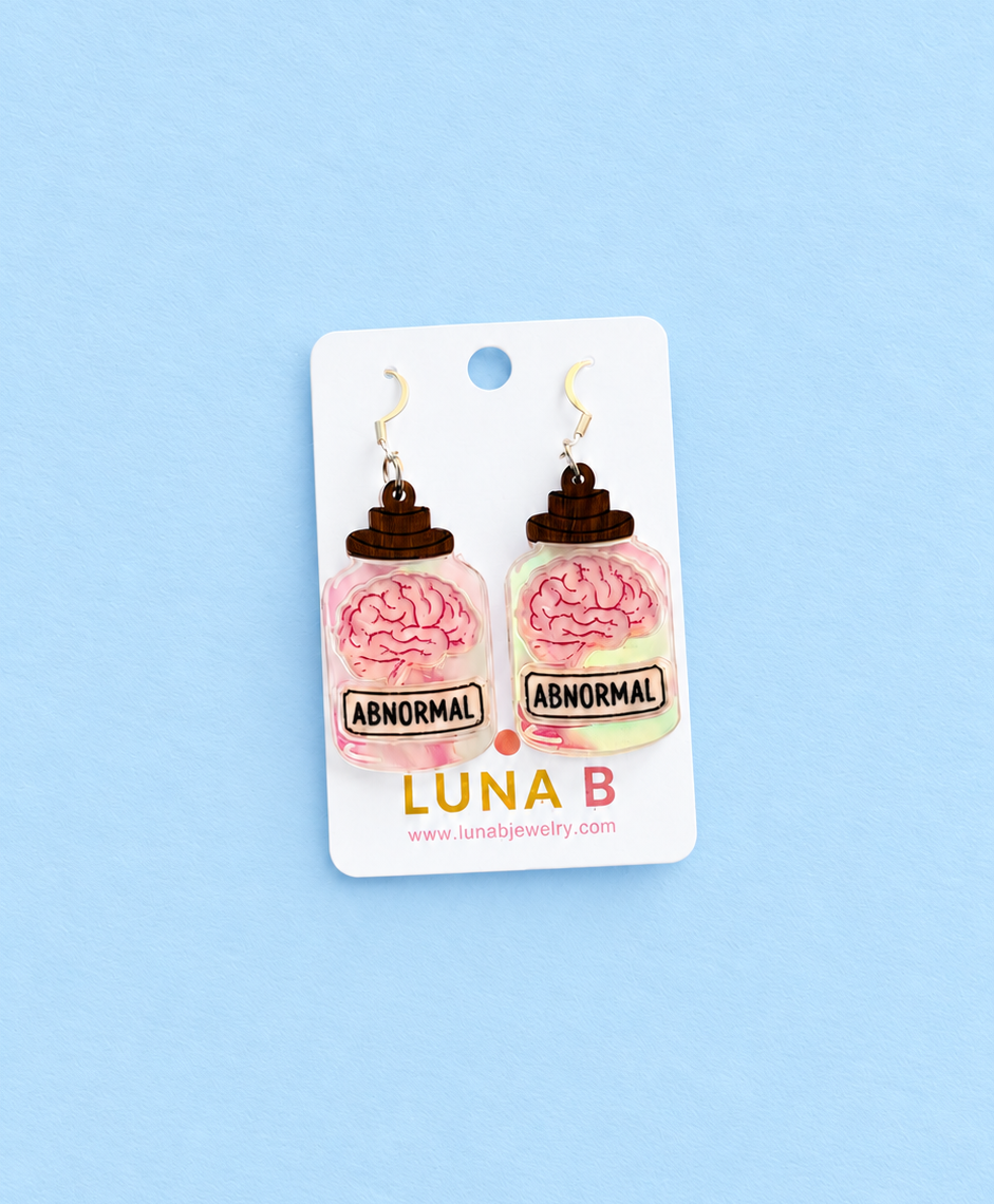 A pair of novelty earrings shaped like pink brain jars labeled 'ABNORMAL' on a white card, displayed against a light blue background. The card shows the brand name 'LUNA B'.