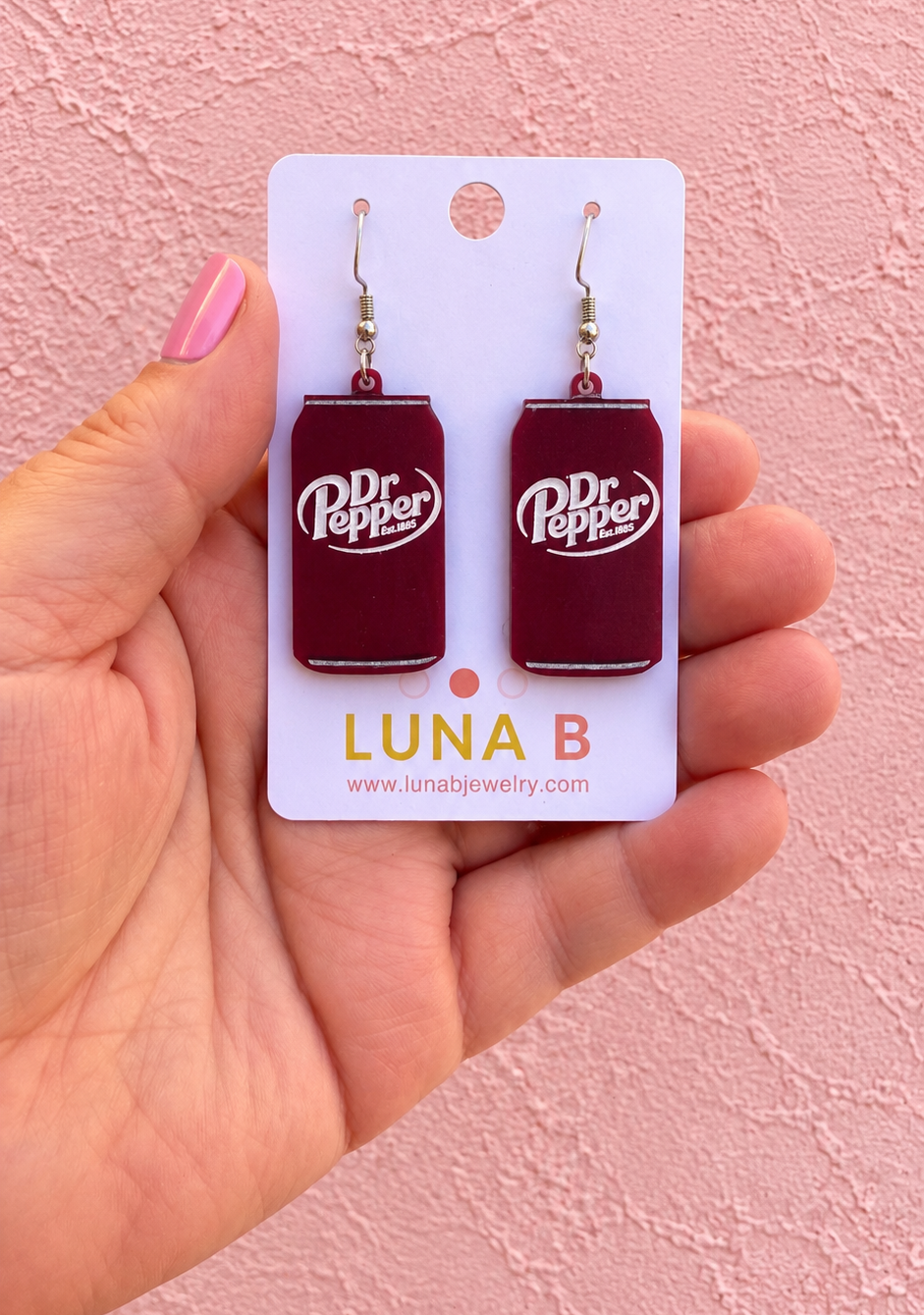 A hand with pink nail polish holds a white card displaying a pair of Dr Pepper can-shaped drop earrings against a textured pink wall.