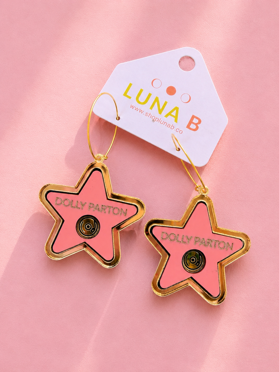A pair of star-shaped enamel pins in pink with gold borders and the text 'DOLLY PARTON' on them, hanging from gold-tone wire hooks from a white brand tag that reads 'LUNA B'. The pins are displayed on a soft pink background.