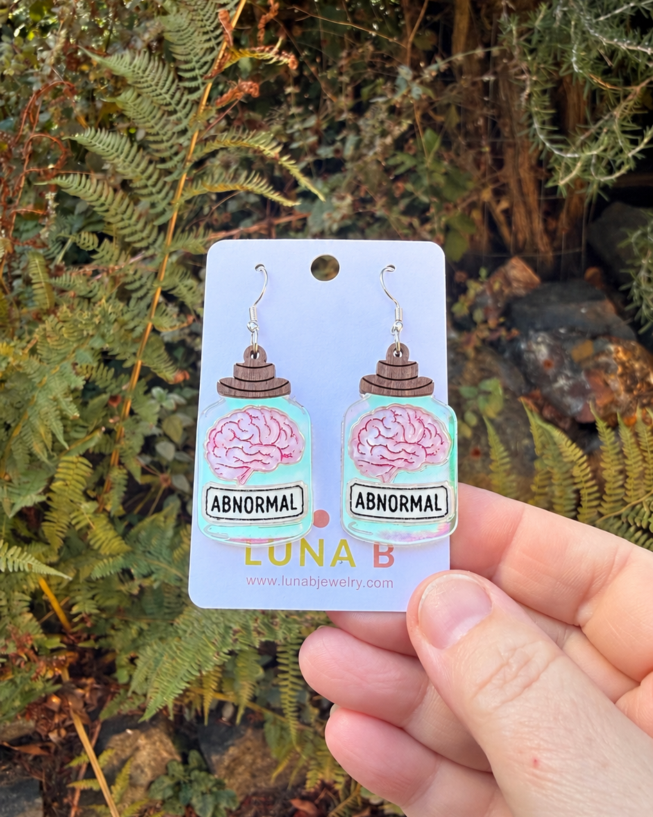 A hand holds a pair of novelty brain-shaped earrings on a white backing card outdoors. The earrings feature a pink brain on a light blue background with a brown cupcake above it and the word 'ABNORMAL' printed below.