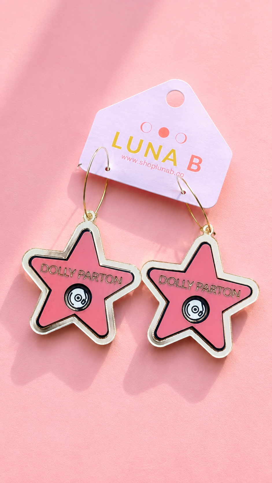 Pink star-shaped enamel earrings with gold-tone hooks and trim, featuring a black-and-white circular center and the text 'DOLLY PARTON' on each star. They hang from a white display card printed with 'LUNA B' against a soft pink background.