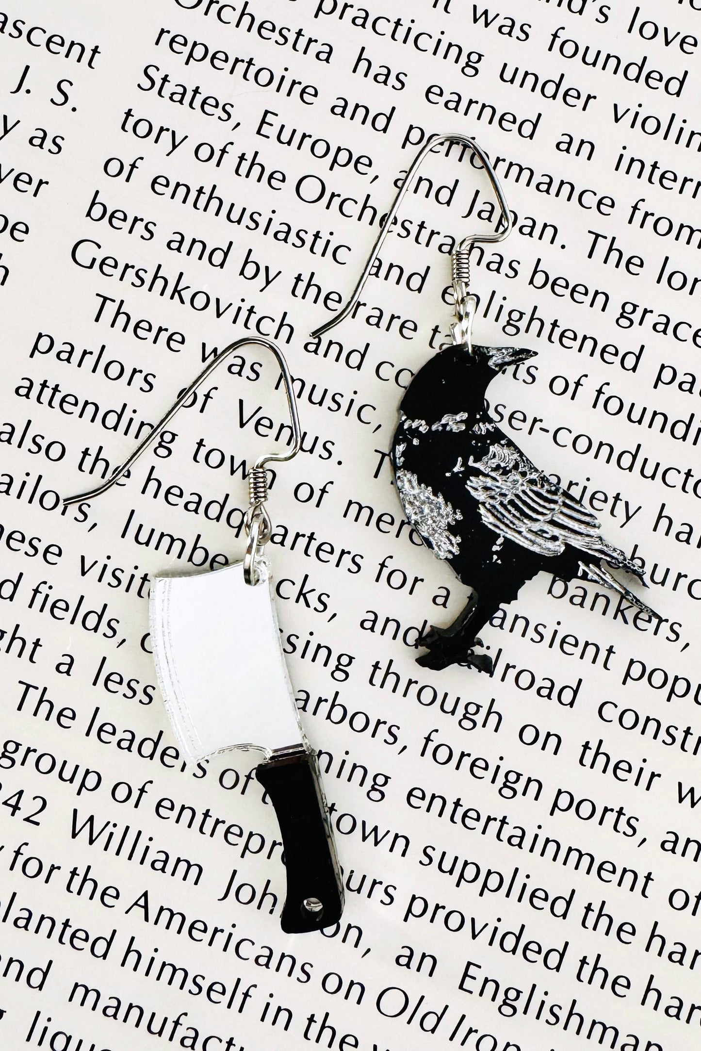 Handmade mismatched statement earrings with a silver butcher knife charm and a black bird charm on stainless steel ear wires, styled on a vintage book page.