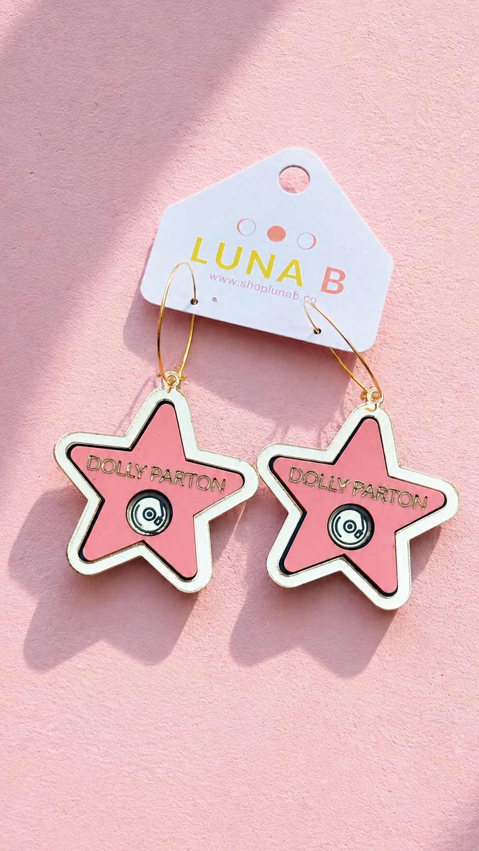 A pair of star-shaped dangle earrings with pink centers, white borders, and gold-tone hoops, hanging from a white display card that reads 'LUNA B'. The card is placed on a pink background.