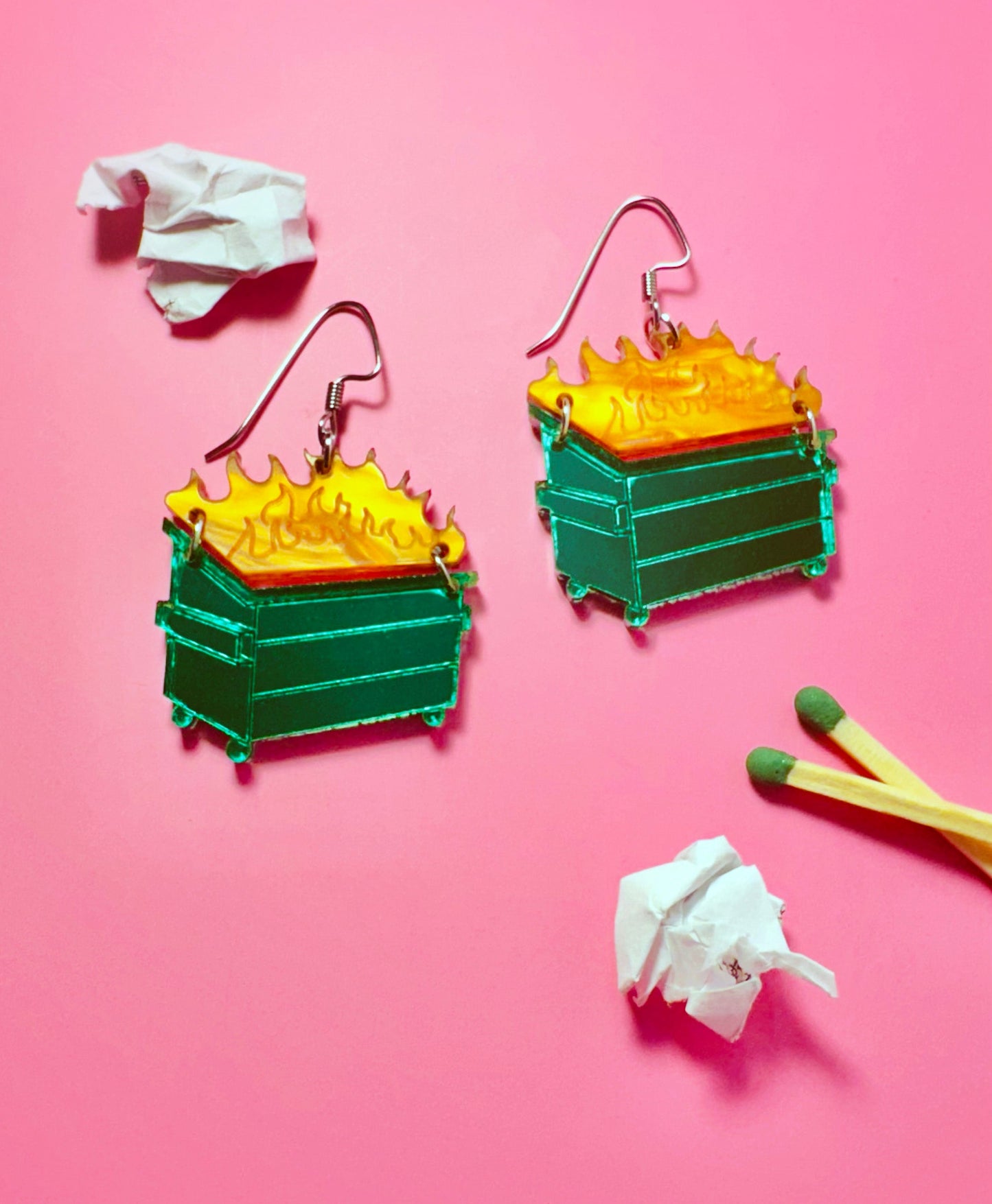 Colorful earrings resembling green trash cans with yellow flames on a pink background.