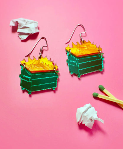 Colorful earrings resembling green trash cans with yellow flames on a pink background.