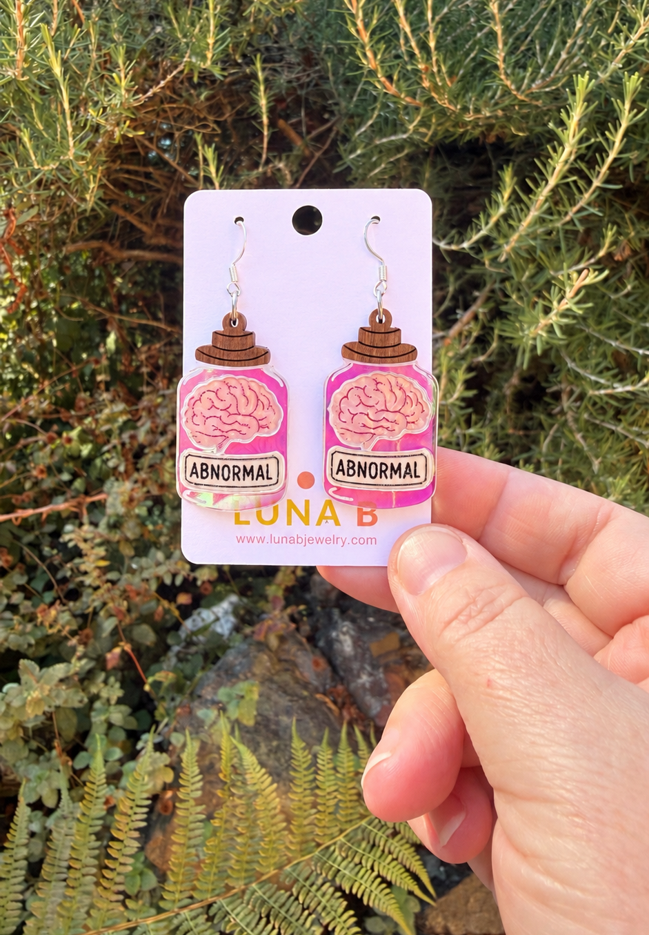 A hand holds a white display card featuring a pair of dangling earrings shaped like pink brain jars labeled 'ABNORMAL.' The background is an outdoor garden with green foliage.