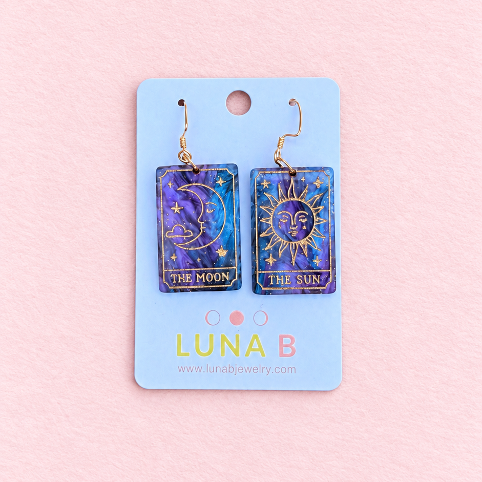Mismatched Sun and Moon tarot card earrings made from swirling purple and midnight blue acrylic, featuring gold engraved celestial illustrations and gold-tone hooks, displayed on a Luna B Jewelry card against a soft pink background.