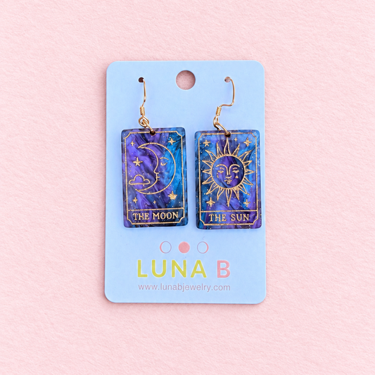Mismatched Sun and Moon tarot card earrings made from swirling purple and midnight blue acrylic, featuring gold engraved celestial illustrations and gold-tone hooks, displayed on a Luna B Jewelry card against a soft pink background.