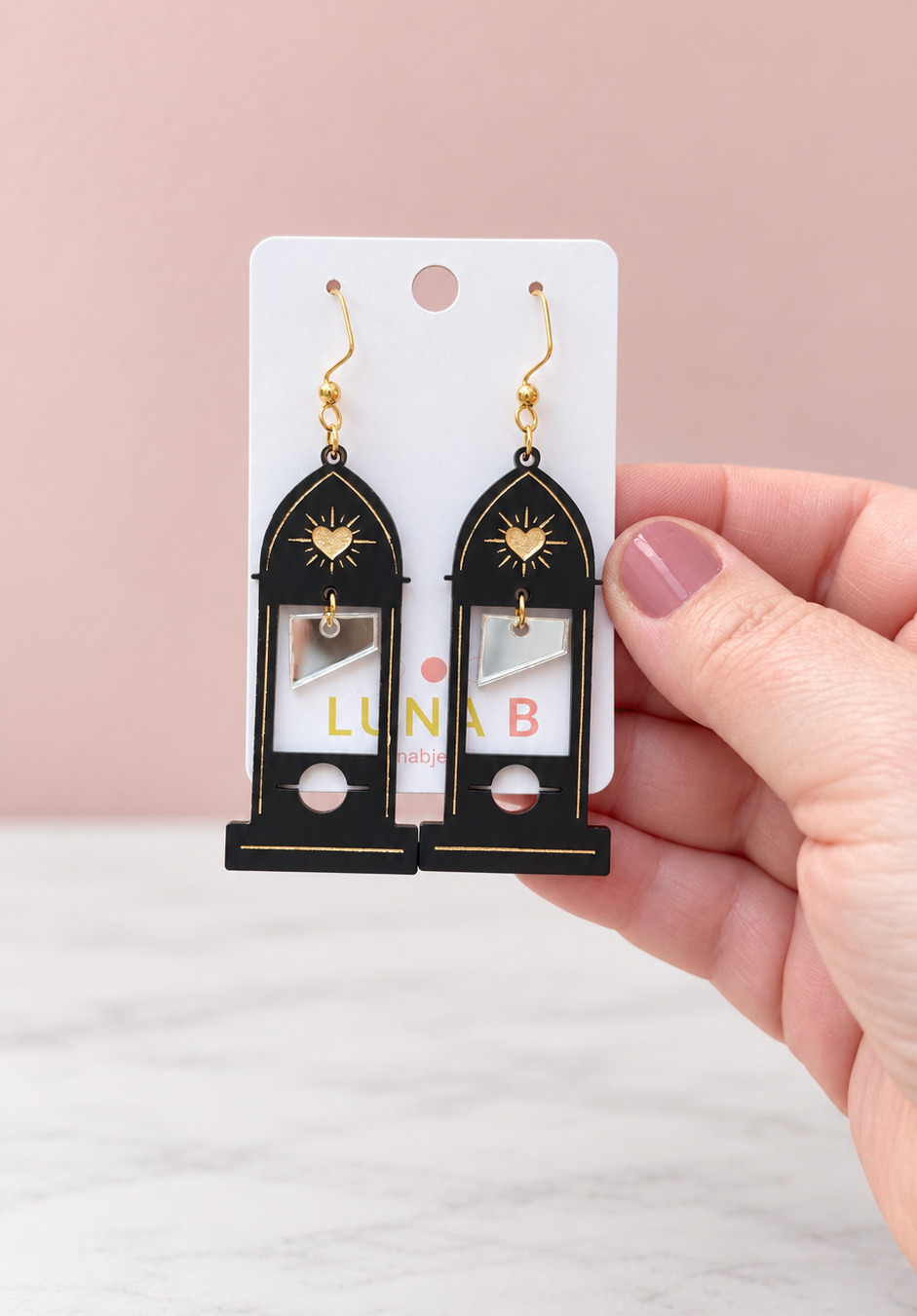 Black acrylic guillotine earrings with pointed Gothic cathedral arches, gold sacred heart accents, and dangling silver mirror blades, displayed on a Luna B Jewelry card against a soft pink background.