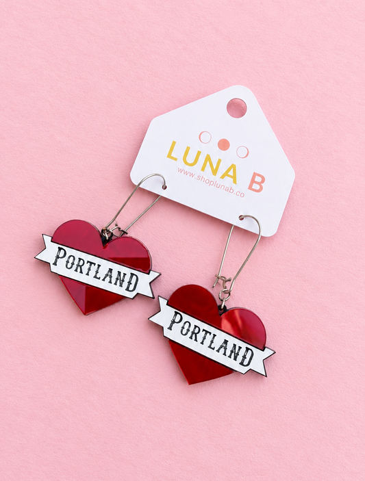 A pair of red heart-shaped drop earrings with white banner charms that read 'PORTLAND' in black lettering, hanging from silver hooks from a white branded display card that says 'LUNA B'. The earrings are photographed on a solid light pink background.