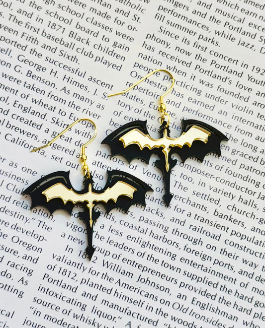 Black acrylic dragon silhouette earrings with gold detailing on stainless steel hooks, photographed on an open book page.