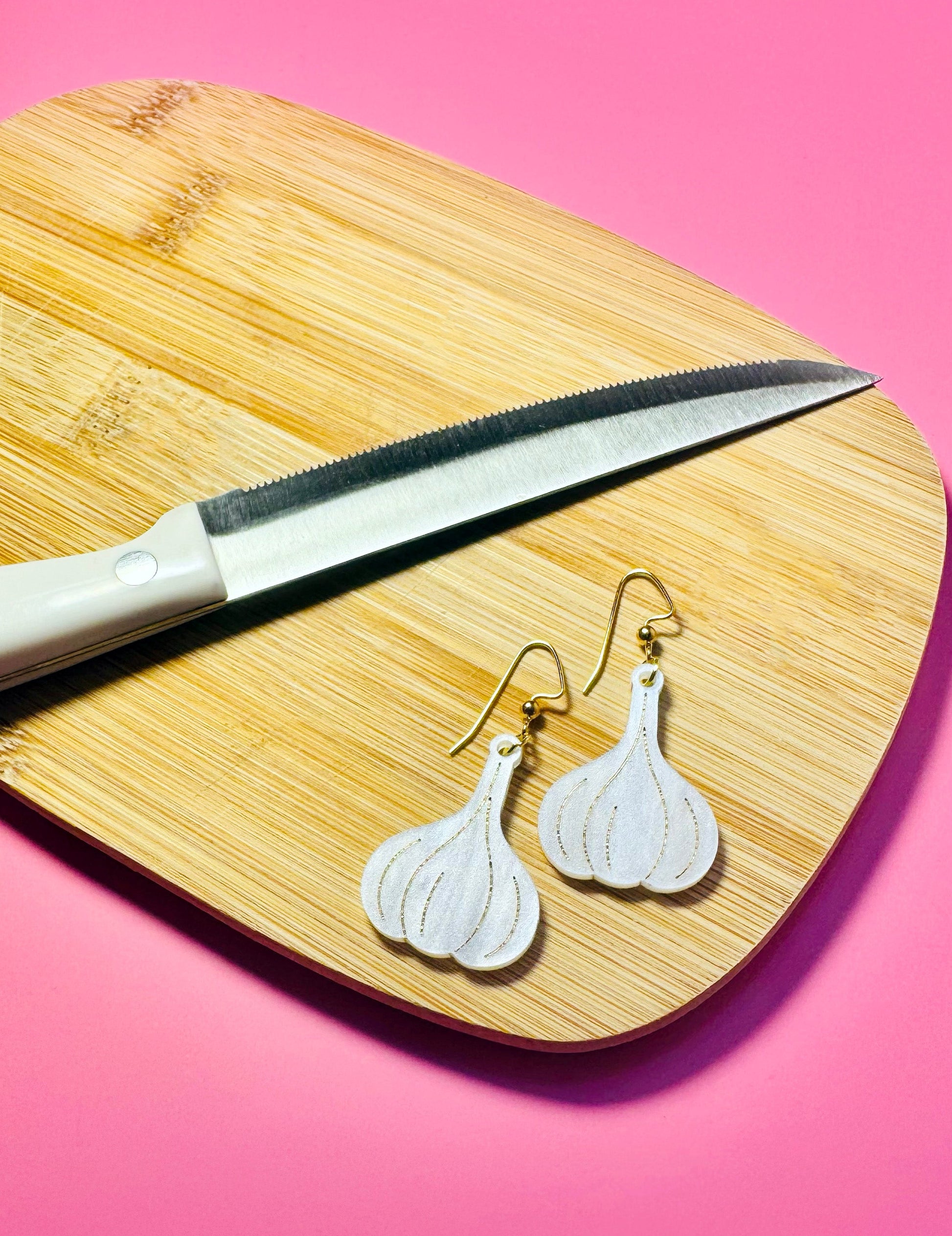Garlic-shaped earrings on a wooden cutting board with a knife against a pink background