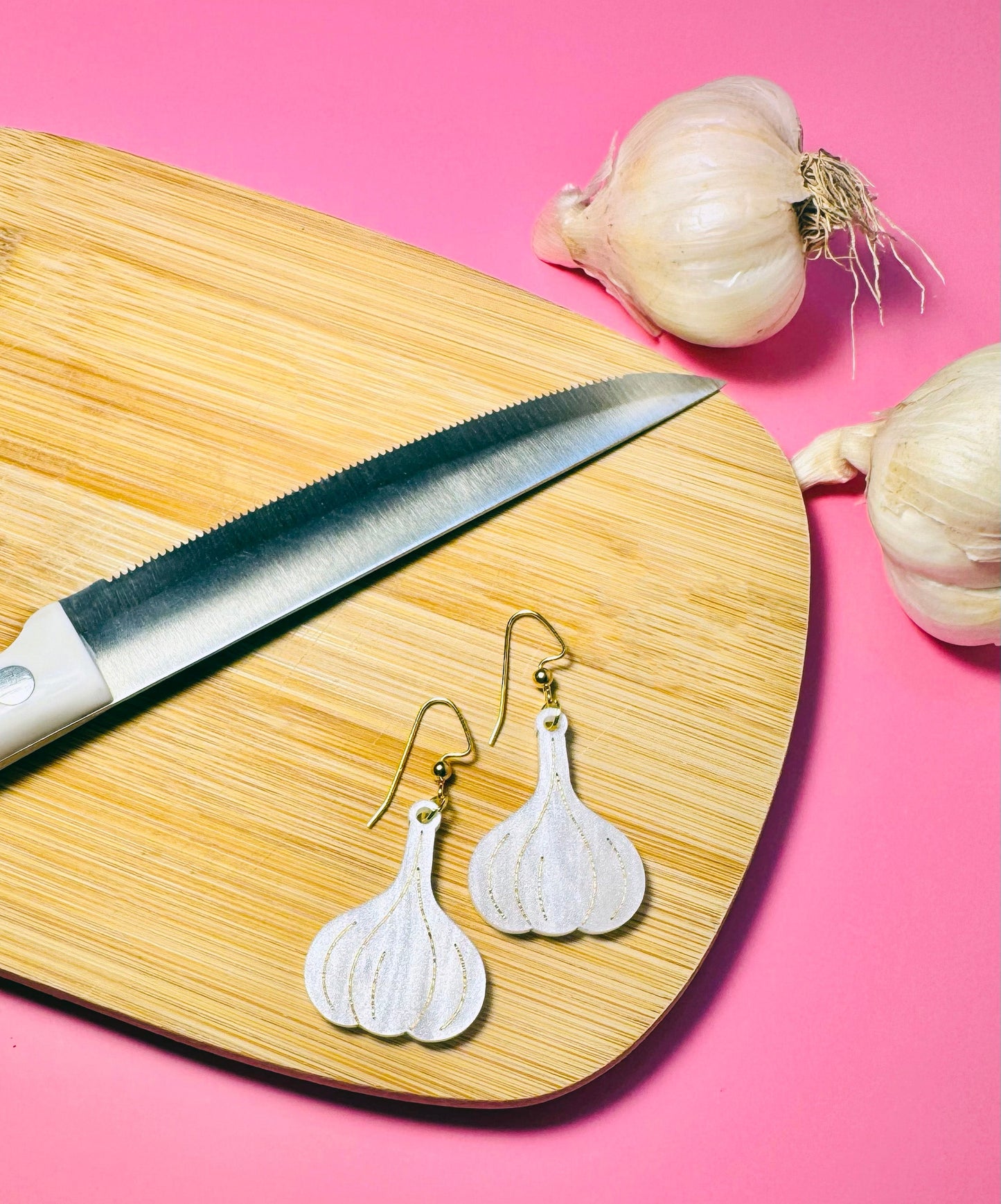Garlic-shaped earrings on a wooden cutting board with a knife and garlic bulbs against a pink background