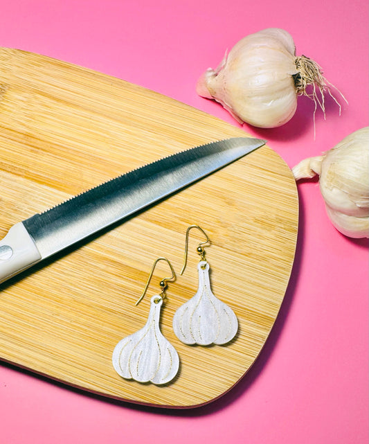 Garlic-shaped earrings on a wooden cutting board with a knife and garlic bulbs against a pink background