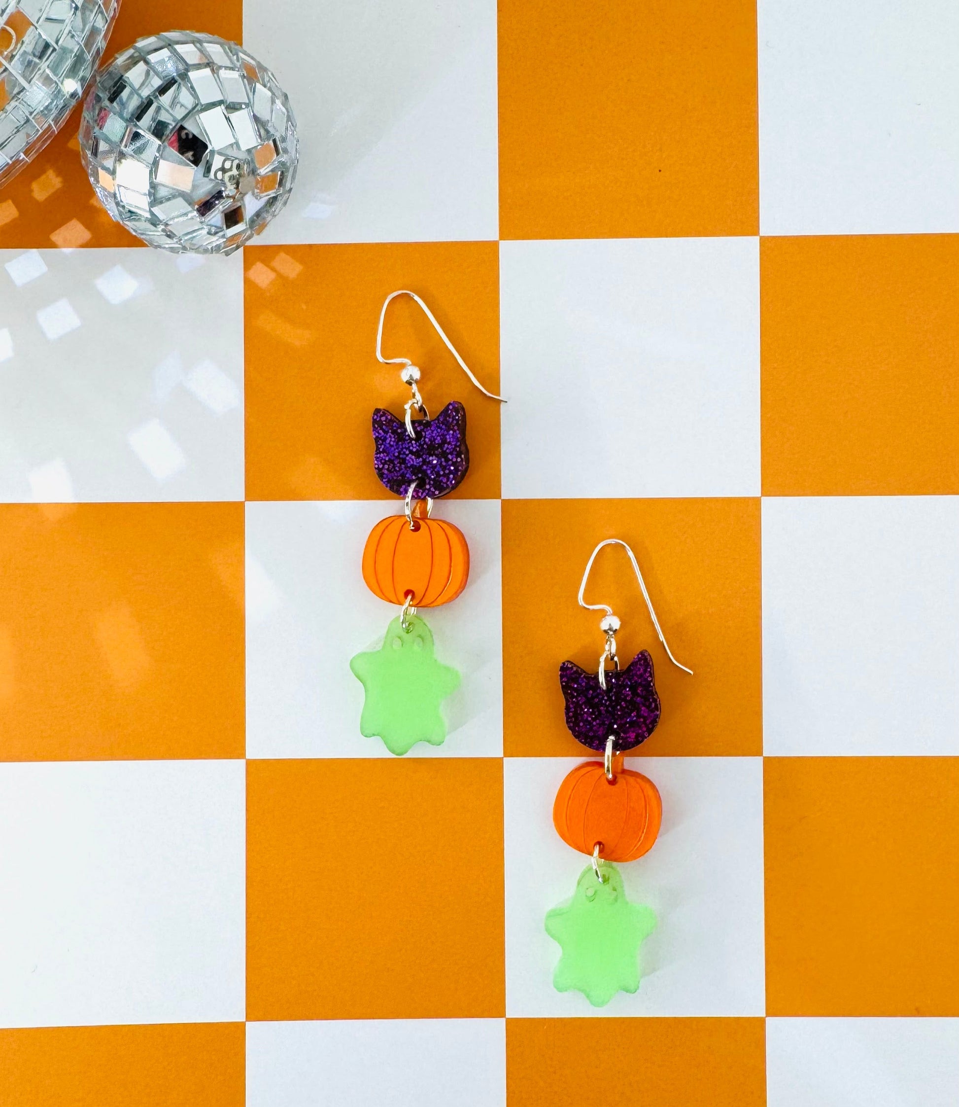 Halloween-themed earrings on a checkered orange and white surface with a disco ball.