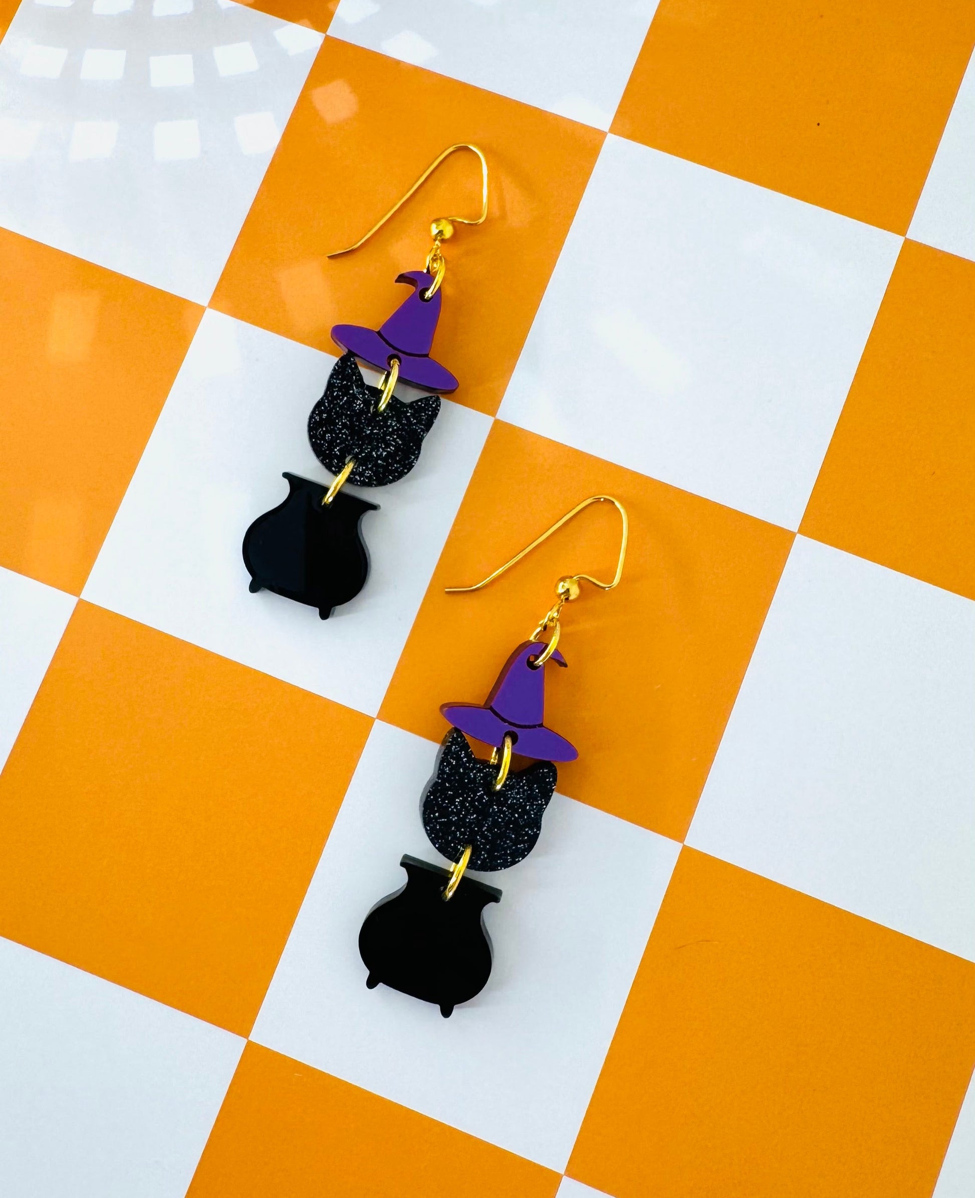 Black cat-shaped earrings with purple hats on a checkered orange and white surface.