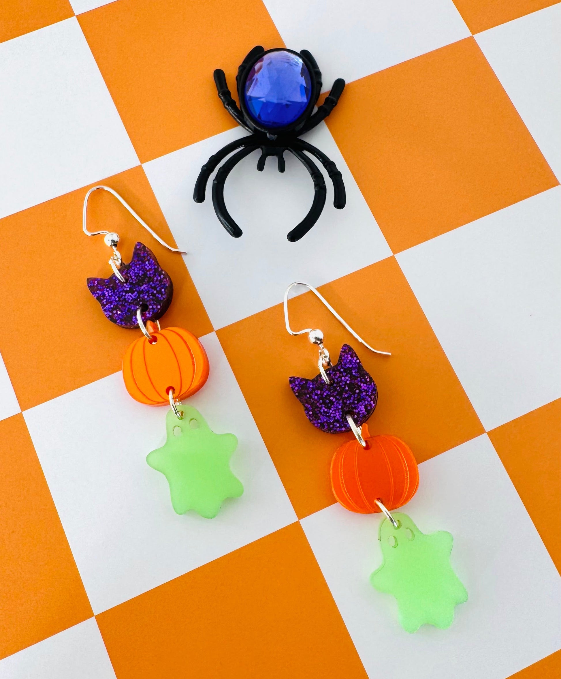 Halloween-themed earrings with pumpkins, ghosts, and a spider on a checkered background.
