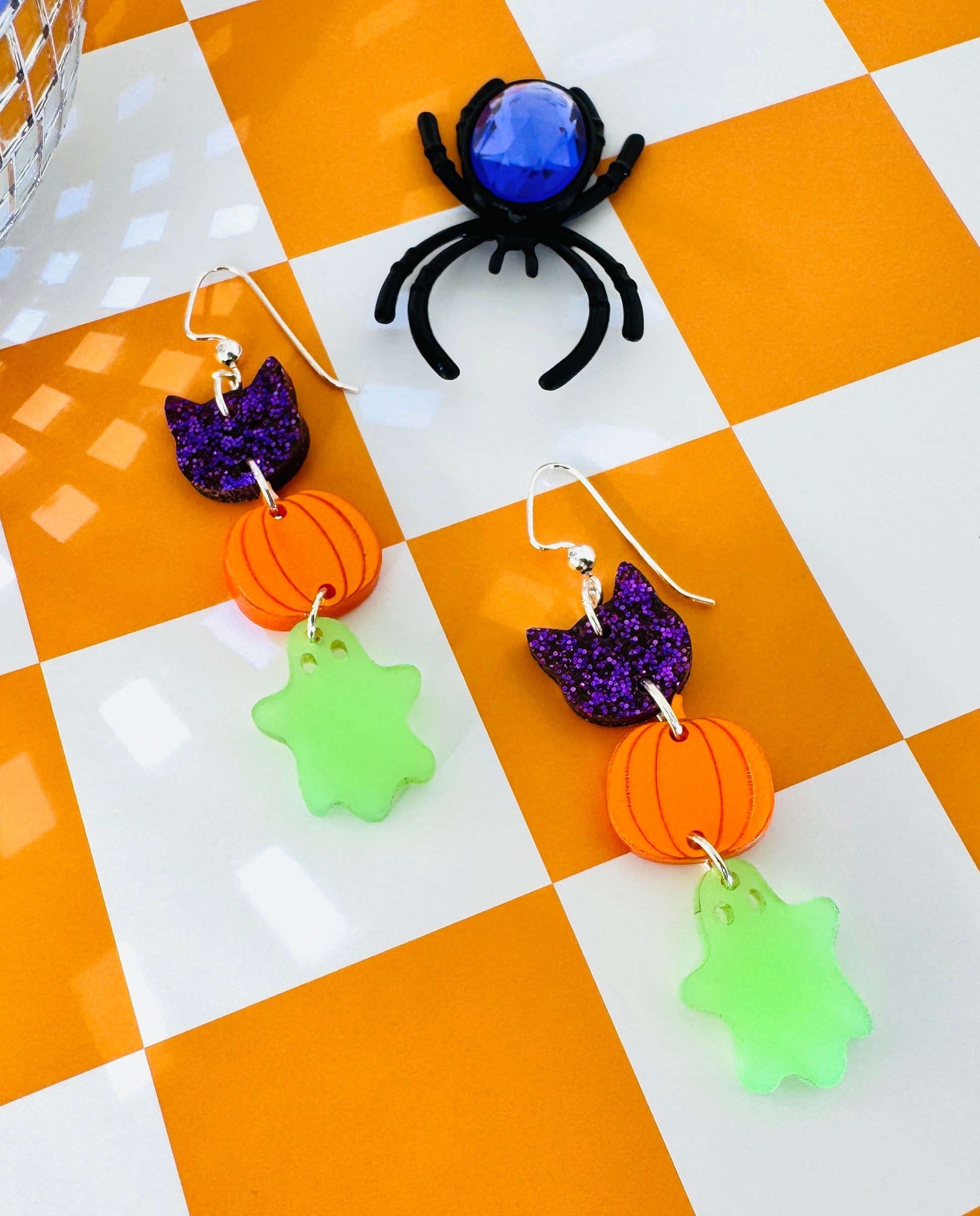 Halloween-themed earrings with pumpkins, ghosts, and a spider on a checkered background.