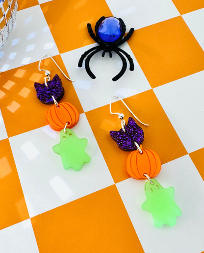 Halloween-themed earrings with pumpkins, ghosts, and a spider on a checkered background.