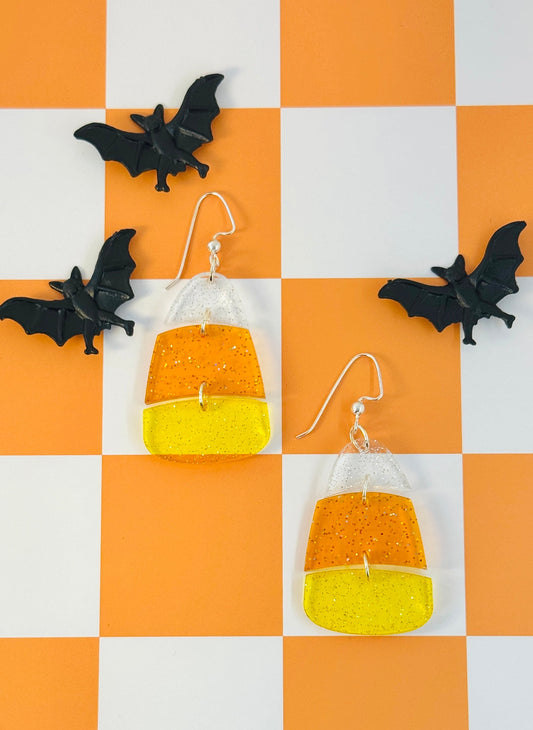 Halloween-themed candy corn earrings on a checkered orange and white background with small black bats.