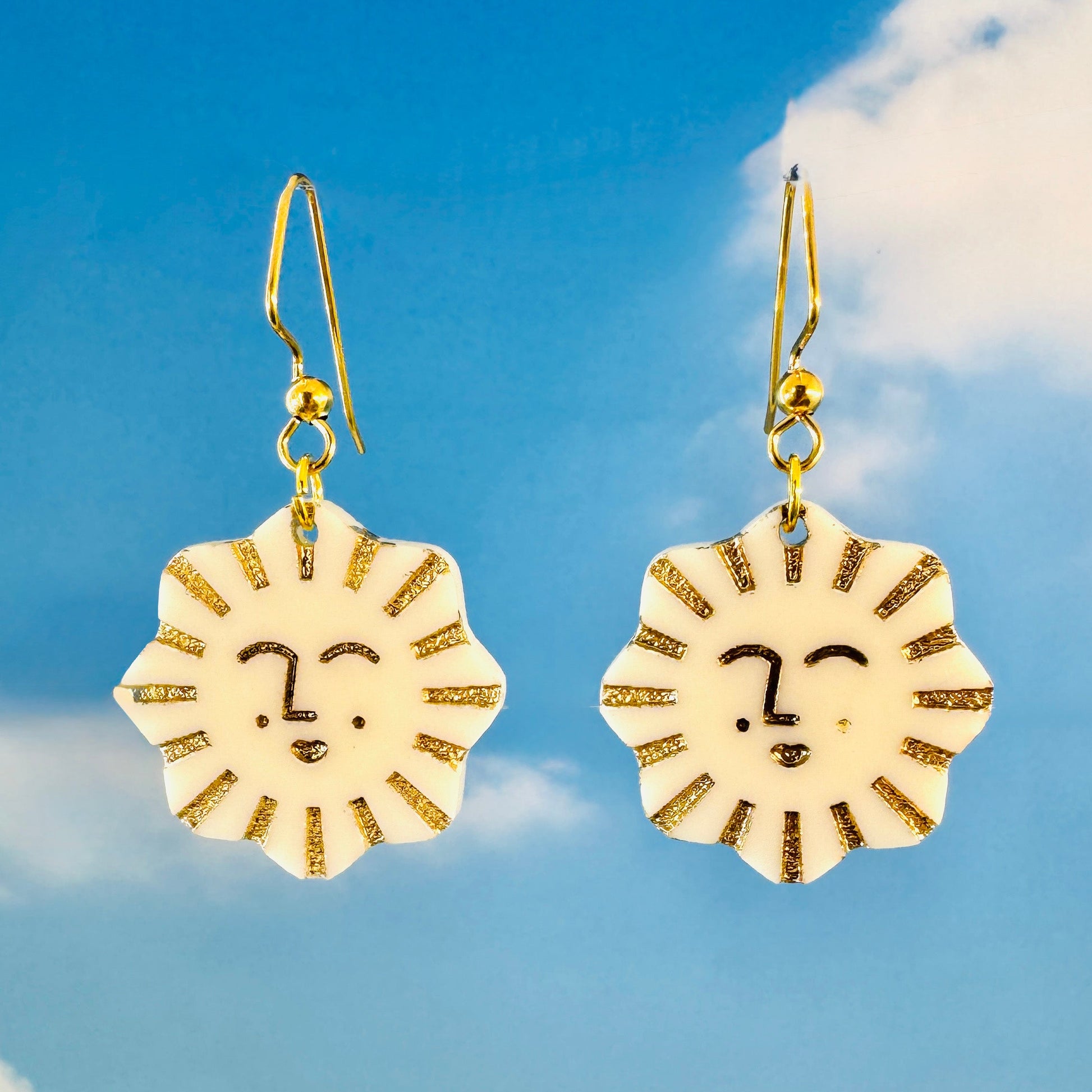 Gold sun-shaped earrings with faces against a blue sky background