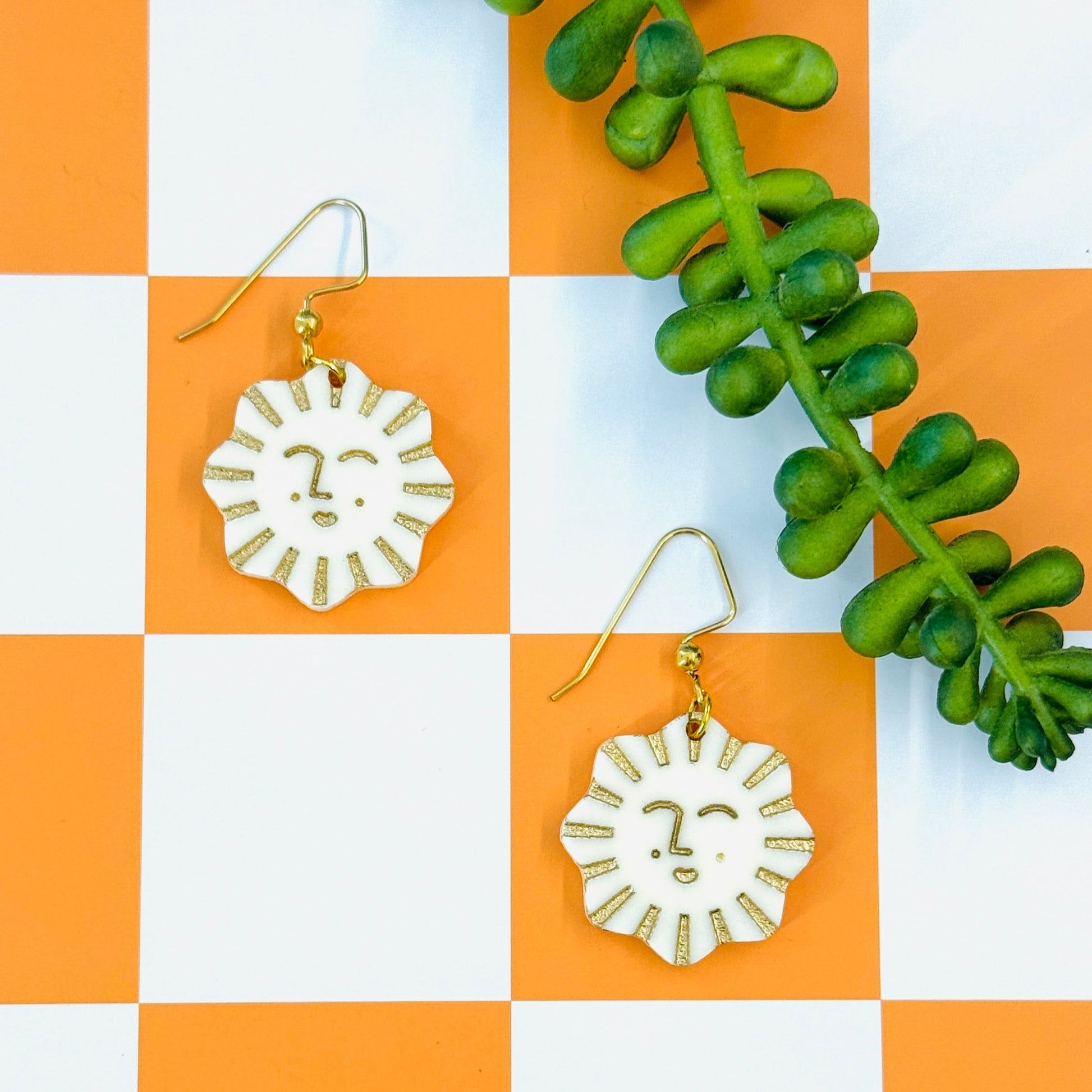 Sun-shaped earrings on a checkered orange and white background with green leaves.