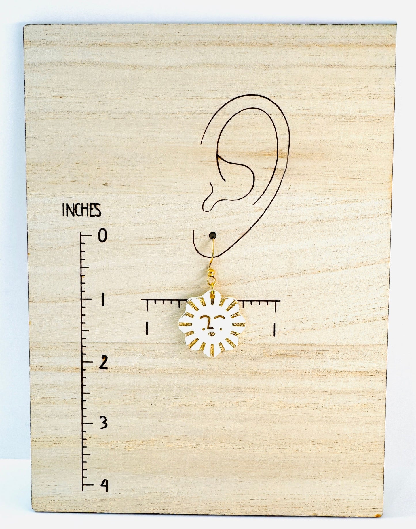 Gold sun-shaped earring on a wooden block with an ear illustration and ruler for scale.